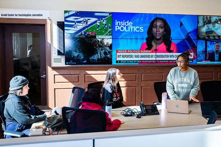 A group of students works on laptops in front of several large monitors displaying an array of news stories.